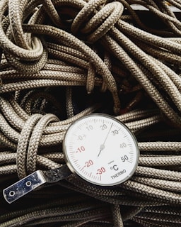 A close-up view of a thermometer resting on a pile of coiled ropes. The thermometer displays a range of temperatures in degrees Celsius, with prominent numbers and a needle indicator pointing upwards. The ropes have a detailed braided texture and are lightly colored, providing contrast with the metallic and white surface of the thermometer.