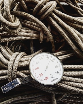 A close-up view of a thermometer resting on a pile of coiled ropes. The thermometer displays a range of temperatures in degrees Celsius, with prominent numbers and a needle indicator pointing upwards. The ropes have a detailed braided texture and are lightly colored, providing contrast with the metallic and white surface of the thermometer.