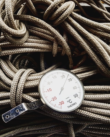A close-up view of a thermometer resting on a pile of coiled ropes. The thermometer displays a range of temperatures in degrees Celsius, with prominent numbers and a needle indicator pointing upwards. The ropes have a detailed braided texture and are lightly colored, providing contrast with the metallic and white surface of the thermometer.