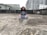 A person is seated cross-legged on a rooftop, meditating with eyes closed. The rooftop has a weathered, concrete surface and is surrounded by high-rise buildings. The sky appears overcast.