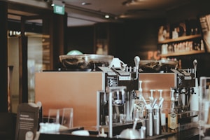 A cozy coffee shop interior featuring two large espresso machines on a counter. Various coffee-making accessories such as cups, milk frothers, and bottles are visible. The background includes shelves with coffee products displayed, and the lighting is warm and ambient.