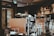 A cozy coffee shop interior featuring two large espresso machines on a counter. Various coffee-making accessories such as cups, milk frothers, and bottles are visible. The background includes shelves with coffee products displayed, and the lighting is warm and ambient.