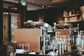 A cozy coffee shop interior featuring two large espresso machines on a counter. Various coffee-making accessories such as cups, milk frothers, and bottles are visible. The background includes shelves with coffee products displayed, and the lighting is warm and ambient.