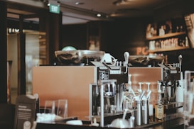A cozy coffee shop interior featuring two large espresso machines on a counter. Various coffee-making accessories such as cups, milk frothers, and bottles are visible. The background includes shelves with coffee products displayed, and the lighting is warm and ambient.
