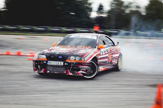 A car decorated with vibrant, multicolored patterns is drifting on a track surrounded by orange traffic cones. Smoke can be seen coming from the rear tires as the car skids, suggesting high speed and precision driving. The environment appears to be an open space or race track with blurred trees and barriers in the background.
