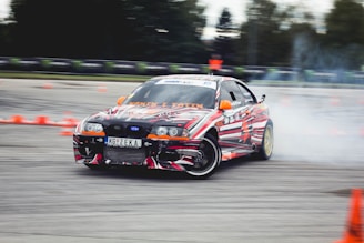 A car decorated with vibrant, multicolored patterns is drifting on a track surrounded by orange traffic cones. Smoke can be seen coming from the rear tires as the car skids, suggesting high speed and precision driving. The environment appears to be an open space or race track with blurred trees and barriers in the background.