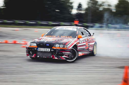 A car decorated with vibrant, multicolored patterns is drifting on a track surrounded by orange traffic cones. Smoke can be seen coming from the rear tires as the car skids, suggesting high speed and precision driving. The environment appears to be an open space or race track with blurred trees and barriers in the background.