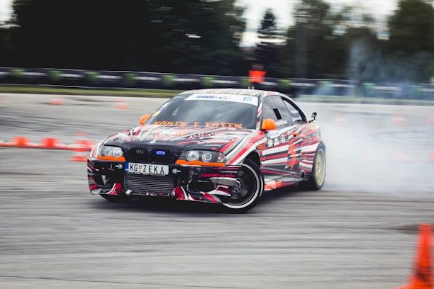 A car decorated with vibrant, multicolored patterns is drifting on a track surrounded by orange traffic cones. Smoke can be seen coming from the rear tires as the car skids, suggesting high speed and precision driving. The environment appears to be an open space or race track with blurred trees and barriers in the background.