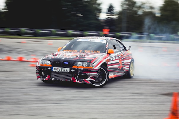 A car decorated with vibrant, multicolored patterns is drifting on a track surrounded by orange traffic cones. Smoke can be seen coming from the rear tires as the car skids, suggesting high speed and precision driving. The environment appears to be an open space or race track with blurred trees and barriers in the background.