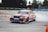 A car decorated with vibrant, multicolored patterns is drifting on a track surrounded by orange traffic cones. Smoke can be seen coming from the rear tires as the car skids, suggesting high speed and precision driving. The environment appears to be an open space or race track with blurred trees and barriers in the background.