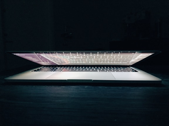 A partially closed laptop sits on a dark surface, with the screen displaying text or code that casts a soft glow. The keyboard is visible, and the edges of the screen are illuminated against the dark background.