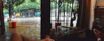 A rainy street scene viewed from inside a cafe or restaurant. Two cups, a lighter, and small items are on a table near an open window with metal bars. Outside, a few motorbikes are parked, and trees line the sidewalk. The pavement glistens with rain, creating a calm urban atmosphere.