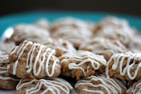 A pile of freshly baked cookies drizzled with white icing, placed on a teal plate. The cookies appear to be oatmeal-based with a textured surface, and the icing is artistically applied in an irregular pattern.