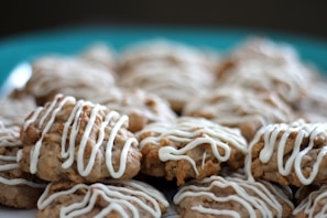 A pile of freshly baked cookies drizzled with white icing, placed on a teal plate. The cookies appear to be oatmeal-based with a textured surface, and the icing is artistically applied in an irregular pattern.