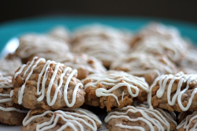 A pile of freshly baked cookies drizzled with white icing, placed on a teal plate. The cookies appear to be oatmeal-based with a textured surface, and the icing is artistically applied in an irregular pattern.