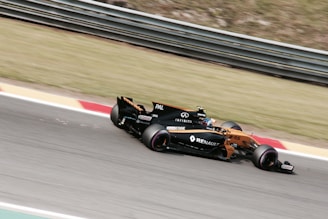 A Formula 1 car is racing on a track, featuring visible branding and logos such as Renault. The vehicle is painted in a combination of orange and black, and the image captures the motion and speed of the car. The background consists of grass and track barriers, slightly blurred due to the car's movement.
