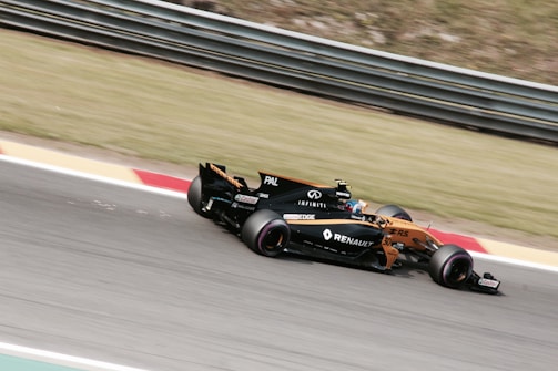 A Formula 1 car is racing on a track, featuring visible branding and logos such as Renault. The vehicle is painted in a combination of orange and black, and the image captures the motion and speed of the car. The background consists of grass and track barriers, slightly blurred due to the car's movement.
