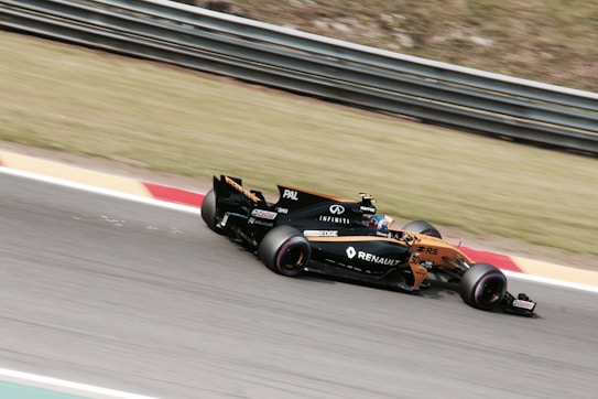 A Formula 1 car is racing on a track, featuring visible branding and logos such as Renault. The vehicle is painted in a combination of orange and black, and the image captures the motion and speed of the car. The background consists of grass and track barriers, slightly blurred due to the car's movement.