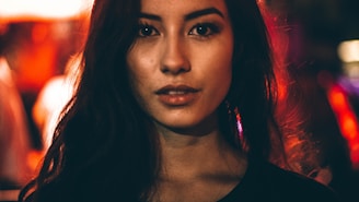 A close-up portrait of a person with long, dark hair, intense eyes, and a neutral expression. The background features soft, blurred, warm-toned lights creating a bokeh effect, suggesting an evening or nighttime setting.