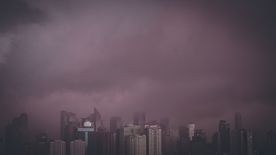 A dramatic cityscape view featuring a skyline filled with towering buildings under a dark, ominous sky filled with storm clouds. The atmosphere is moody and tense, as the clouds seem to be descending toward the city.