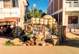 A street scene with a cart displaying an array of handmade baskets and woven items. Colorful textiles hang from the sides, and various woven hats are placed on the ground. The cart is parked between two buildings, and a shop is visible in the background.