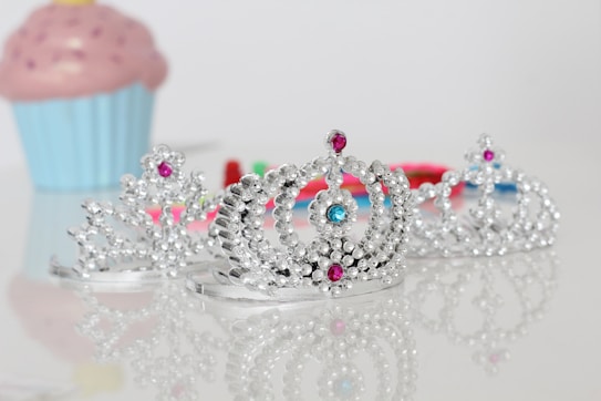 Three sparkling silver tiaras adorned with colorful gems are prominently placed on a reflective surface. In the background, a pastel-colored cupcake adds a playful element. The composition is bright and cheerful, with the focus on the intricate detailing of the crowns.