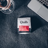 A packet of Quills Coffee labeled 'Inkwell Signature Blend' is placed on a dark textured surface beside a glass coffee brewing device and part of a silver laptop.