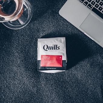 A packet of Quills Coffee labeled 'Inkwell Signature Blend' is placed on a dark textured surface beside a glass coffee brewing device and part of a silver laptop.
