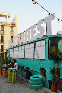 A vintage bus painted in teal is repurposed into a cozy setting, possibly for social gatherings. Large white letters spelling 'BUCHAREST' adorn the roof. Two people are seated at an outdoor table beside the bus, surrounded by potted plants and colorful furniture, creating a relaxed atmosphere.