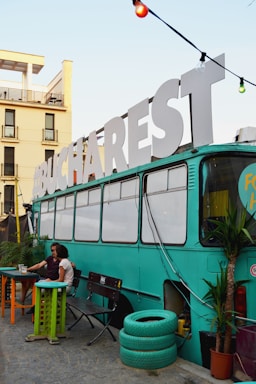 A vintage bus painted in teal is repurposed into a cozy setting, possibly for social gatherings. Large white letters spelling 'BUCHAREST' adorn the roof. Two people are seated at an outdoor table beside the bus, surrounded by potted plants and colorful furniture, creating a relaxed atmosphere.