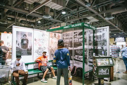 A group of people are gathered around a booth set up to resemble a bus stop, complete with urban street art and graffiti on the walls. There are various posters, including one titled 'The Golden Child'. Two men are seated on a bench, one checking his phone while the other is looking at something outside the frame. A vending machine is nearby, and there are several people in casual clothing walking or standing around. The setting appears to be indoors, possibly at a convention or expo.