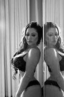 A woman in lace lingerie stands with her eyes closed, leaning against a reflective surface that mirrors her image. The setting has soft lighting and curtains in the background, casting gentle shadows.