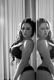 A woman in lace lingerie stands with her eyes closed, leaning against a reflective surface that mirrors her image. The setting has soft lighting and curtains in the background, casting gentle shadows.