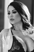 A woman with wavy hair and smokey eye makeup gazes off to the side, wearing a lace bralette and a sheer robe with lace details. She is holding a delicate necklace in a relaxed manner.