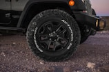 Close-up view of a vehicle's front wheel and tire on a rugged, rocky terrain. The tire is branded with the words 'BFGoodrich All-Terrain T/A' and features a robust tread pattern. Part of the vehicle's front bumper and side body are visible, along with natural elements like stones on the ground.