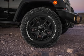 Close-up view of a vehicle's front wheel and tire on a rugged, rocky terrain. The tire is branded with the words 'BFGoodrich All-Terrain T/A' and features a robust tread pattern. Part of the vehicle's front bumper and side body are visible, along with natural elements like stones on the ground.