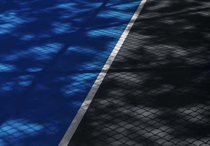 A tennis court surface featuring blue and gray sections divided by a white line. The court is covered with shadows resembling a lattice or net pattern, cast by sunlight filtering through a fence or similar object.