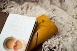A book titled 'The Broken Way' by Ann Voskamp lies on a textured, beige blanket. Next to it is a yellow notebook or journal with an embossed silhouette of a person reaching out to butterflies.