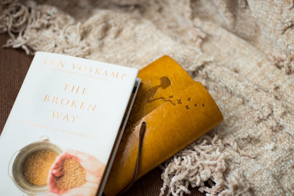 A book titled 'The Broken Way' by Ann Voskamp lies on a textured, beige blanket. Next to it is a yellow notebook or journal with an embossed silhouette of a person reaching out to butterflies.