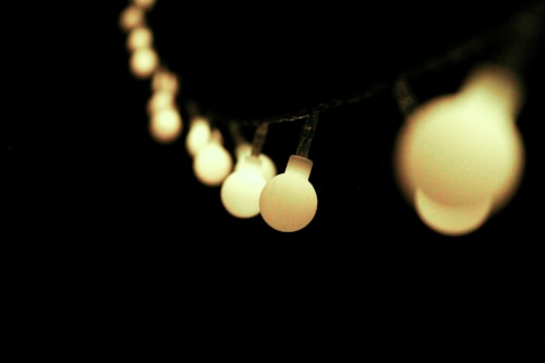 A series of soft, glowing bulb lights are suspended in a line, creating a warm and inviting atmosphere against a dark background. The bulbs have a round shape and emit a gentle, diffused light, enhancing the contrast with the surrounding darkness.