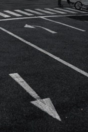 A parking lot with white directional arrows on the asphalt, a pedestrian crosswalk, and part of a bicycle lying on the ground with a glimpse of a person's legs.
