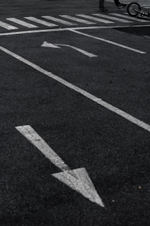 A parking lot with white directional arrows on the asphalt, a pedestrian crosswalk, and part of a bicycle lying on the ground with a glimpse of a person's legs.