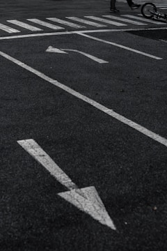 A parking lot with white directional arrows on the asphalt, a pedestrian crosswalk, and part of a bicycle lying on the ground with a glimpse of a person's legs.