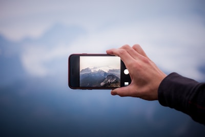 A hand holding a smartphone capturing a photo of a mountainous landscape. The main focus is on the phone screen, which displays the detailed image of the mountains, while the background is slightly blurred.