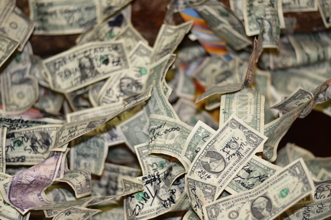 Scattered dollar bills, mainly one-dollar denominations, many of which feature handwritten notes or signatures in black ink, are spread out in an overlapping manner. The background is blurry, creating a sense of depth and emphasizing the focus on the money.