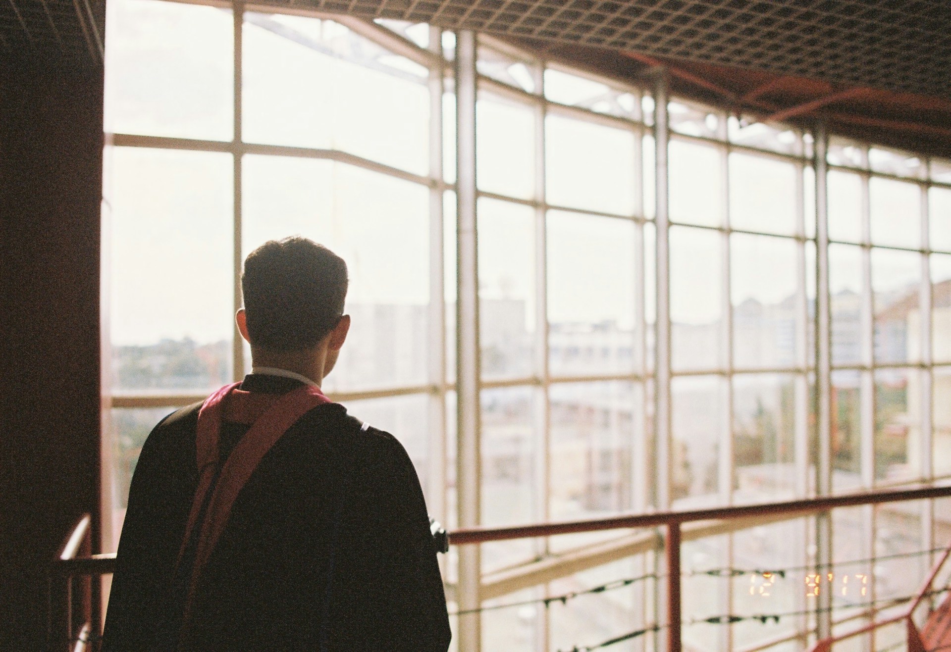 A person in a graduation gown stands with their back to the viewer, looking out through large, grid-like windows. The scene outside appears to be an urban setting, with buildings visible in the background. The lighting is soft, giving a warm glow to the scene.