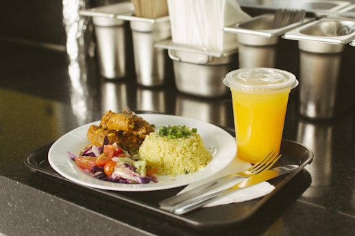 A plate containing rice garnished with chopped herbs, a portion of cooked meat, and a side salad with cherry tomatoes and mixed greens, served with a chilled cup of orange juice on a black tray. The setting appears to be a food service or cafeteria with stainless steel containers in the background.