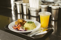 A plate containing rice garnished with chopped herbs, a portion of cooked meat, and a side salad with cherry tomatoes and mixed greens, served with a chilled cup of orange juice on a black tray. The setting appears to be a food service or cafeteria with stainless steel containers in the background.