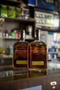 Two bottles of Woodford Reserve bourbon sit on a reflective glass counter in a store. The background features various store shelves with assorted products, slightly out of focus.