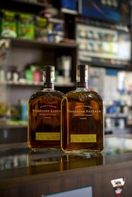 Two bottles of Woodford Reserve bourbon sit on a reflective glass counter in a store. The background features various store shelves with assorted products, slightly out of focus.